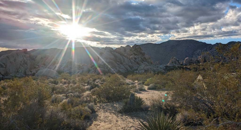 The sun appears just above a mountainous ridge in a desert landscape.